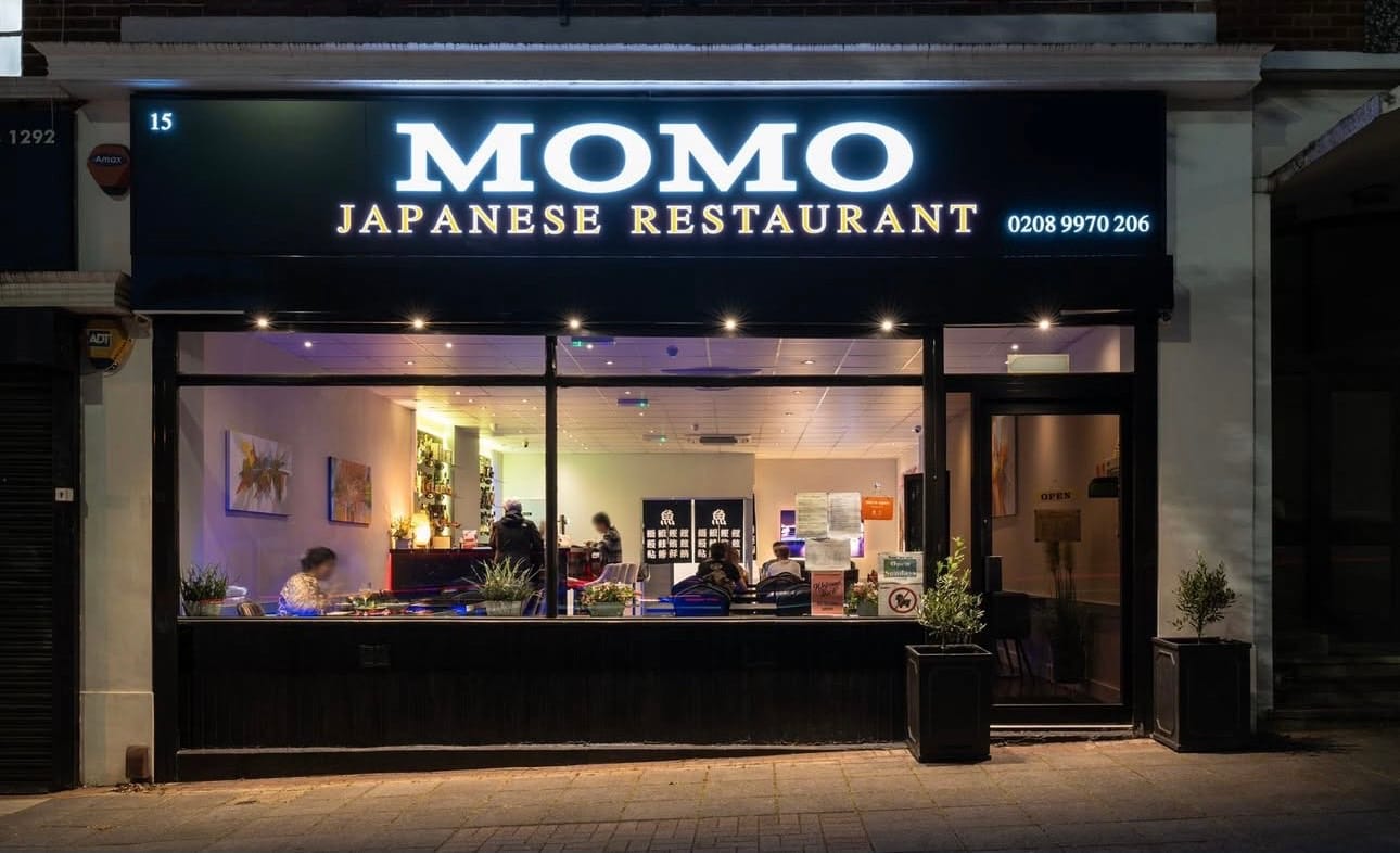 Momo Japanese Restaurant's lit shopfront at night, with the name and phone number above the window, on Ashbourne Parade, Hanger Lane